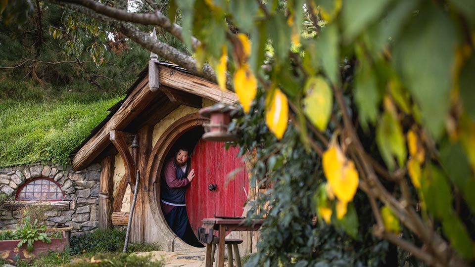 A man peering out of the round red door of a Hobbit house