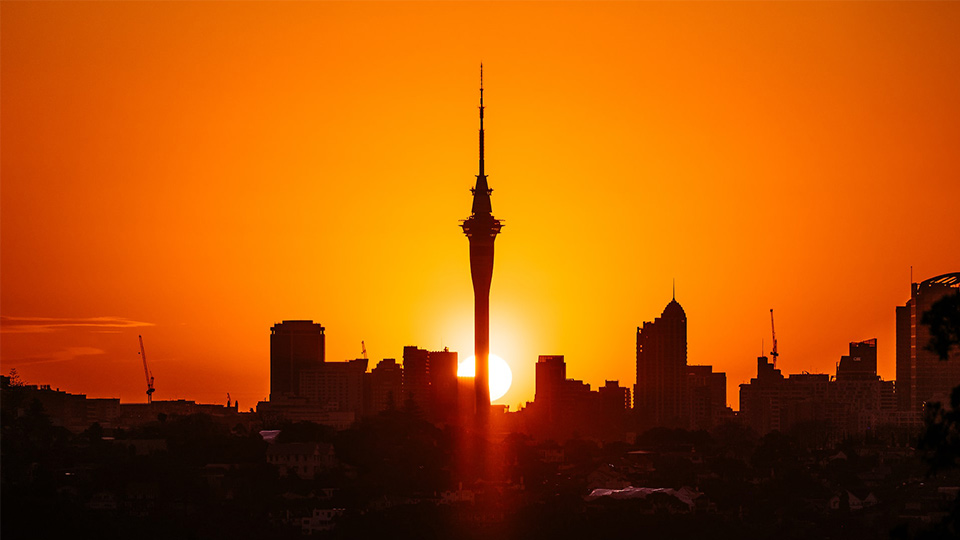 A silhouette of the Auckland skyline in front of the bright orange setting sun with the Sky Tower standing prominently above the other buildings